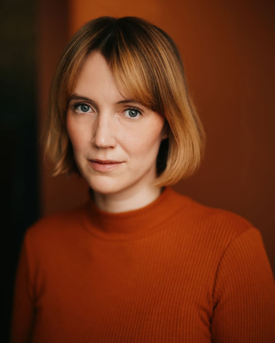 Actor casting headshot of a young woman with auburn hair swept back, blue-grey eyes, and gold hoop earrings, wearing a light grey plaid shirt and looking over her shoulder with a calm, neutral expression against a dark moody background