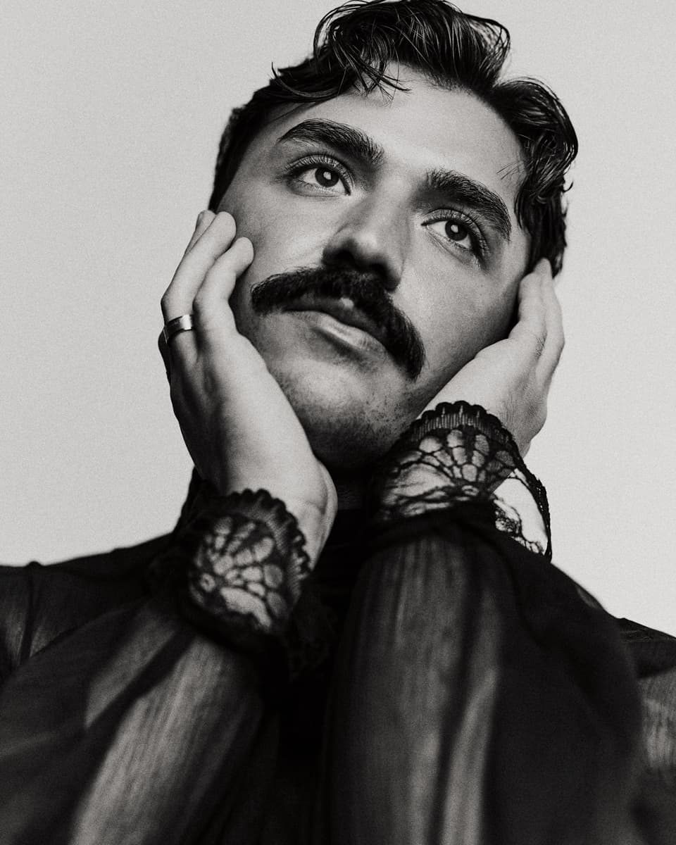 Black and white commercial actor headshot of a man with slicked-back hair and a thick moustache, cradling his face in both hands with lace-cuffed sleeves and a ring, gazing upward with a theatrical expression against a light background