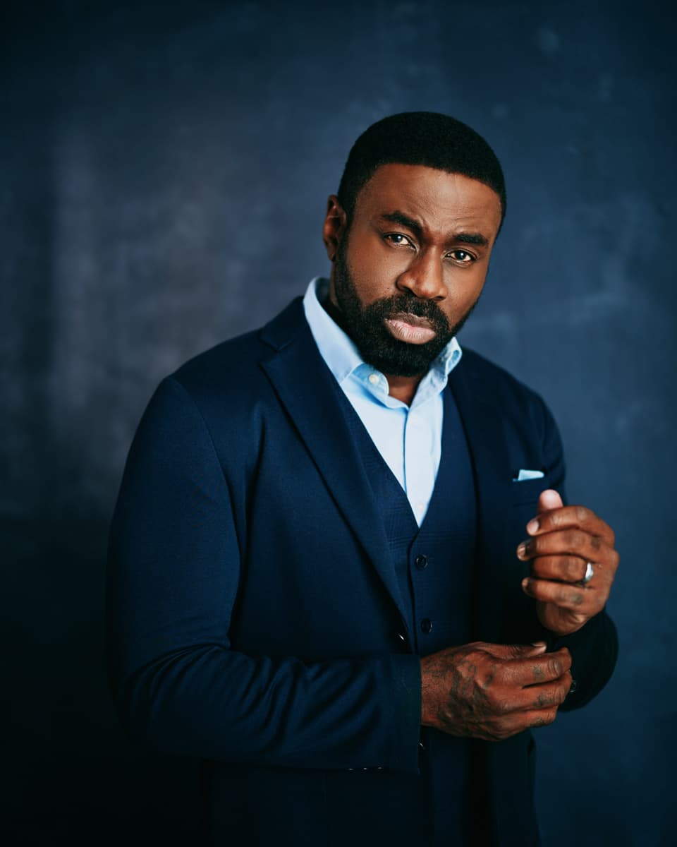 Dramatic actor headshot of a man with a full beard wearing a navy blazer over a light blue shirt, hands clasped, with a commanding and intense gaze against a deep textured blue-black background