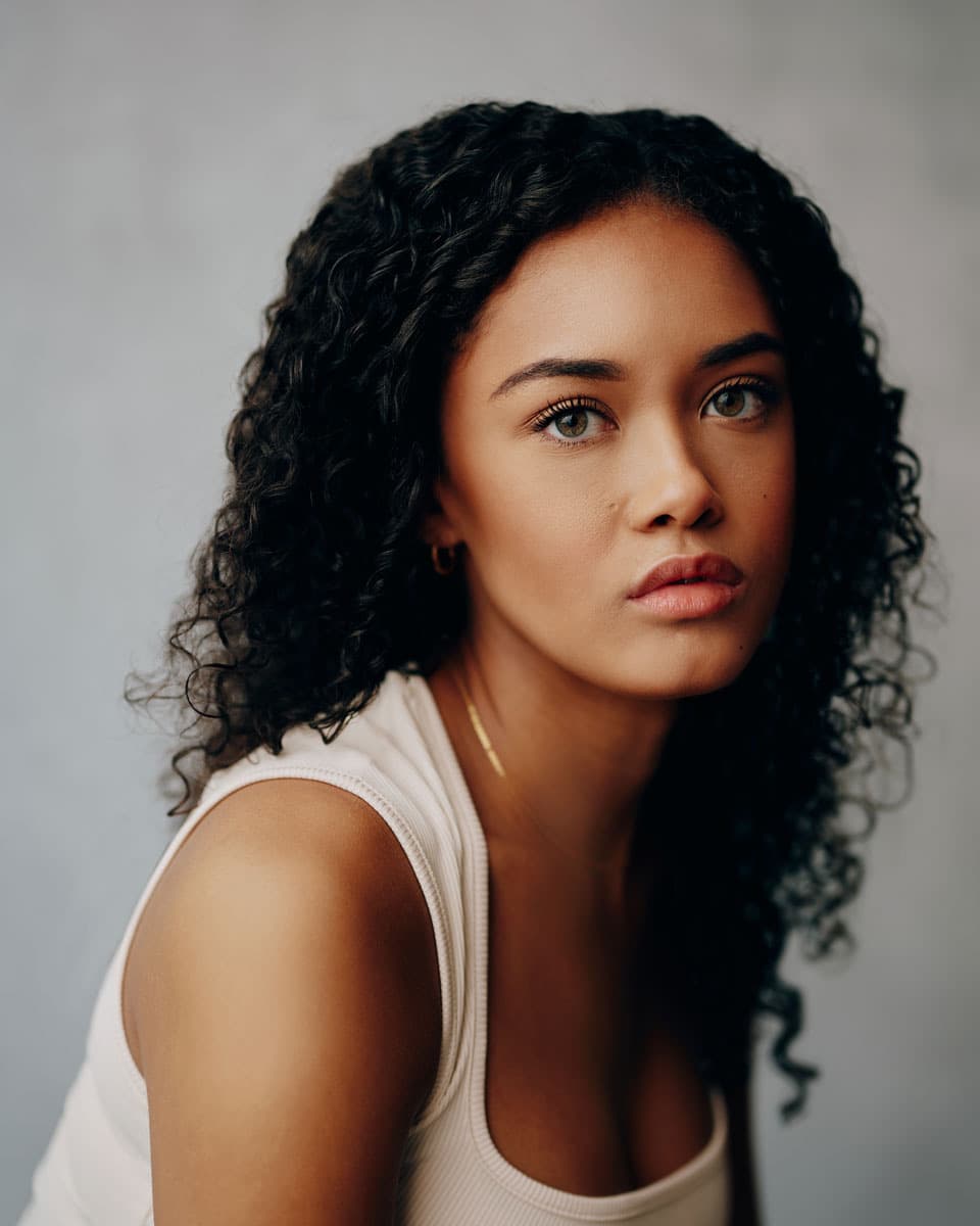 Close-crop actor headshot of a young woman with curly dark hair, gold hoop earrings, and a delicate necklace, wearing a white tank top with a calm, direct gaze against a soft grey backdrop