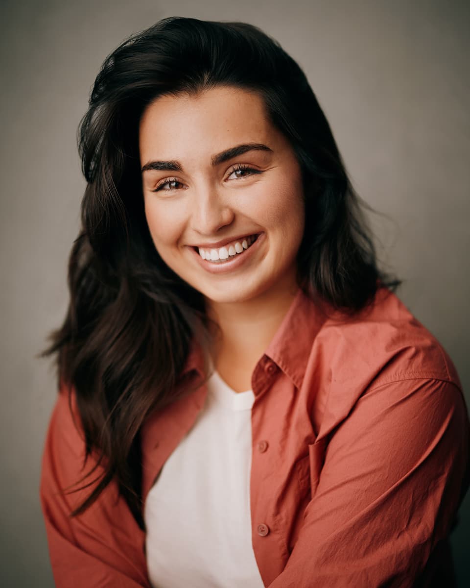 Actor headshot of a young woman with long dark wavy hair wearing a terracotta shirt over a white top, beaming with a wide natural smile against a warm grey backdrop