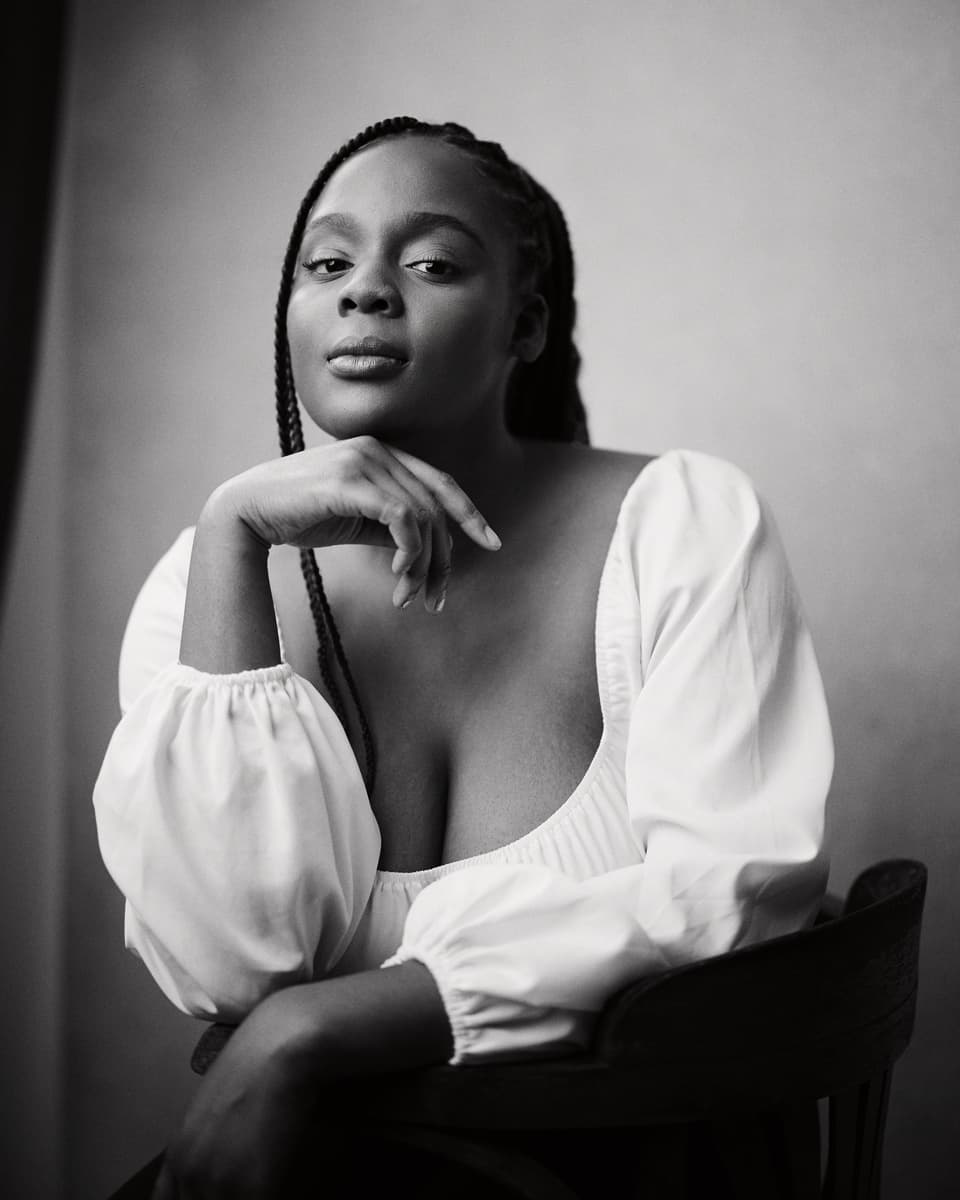 Studio actor headshot of a woman with box braids wearing a white puff-sleeve top, resting her chin on her hand with a serene expression, shot in black and white against a soft light backdrop