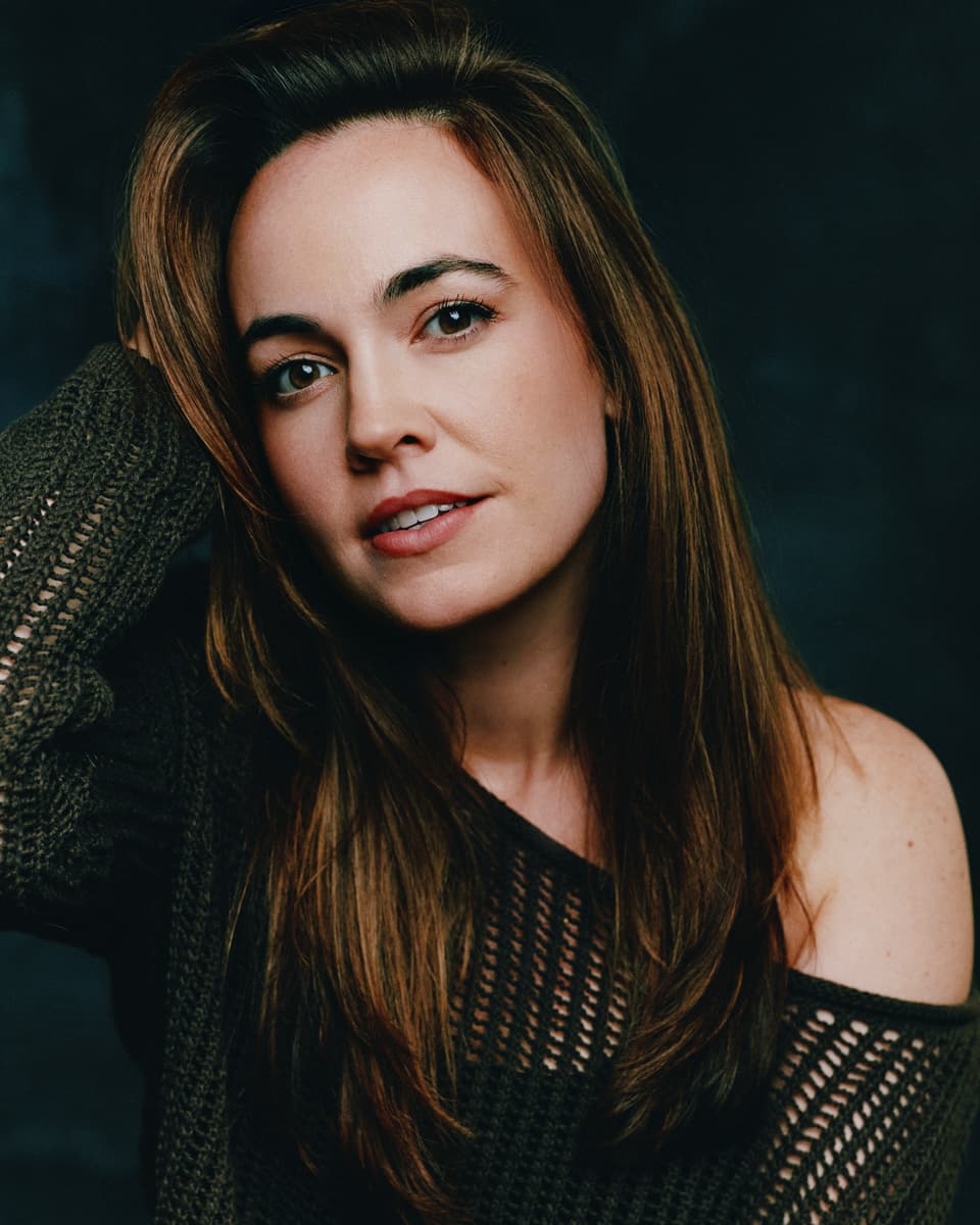 Actress commercial headshot of a woman with long straight brown hair wearing an open-knit off-shoulder dark sweater, hand raised to her head with a relaxed half-smile against a dark moody blue-grey backdrop