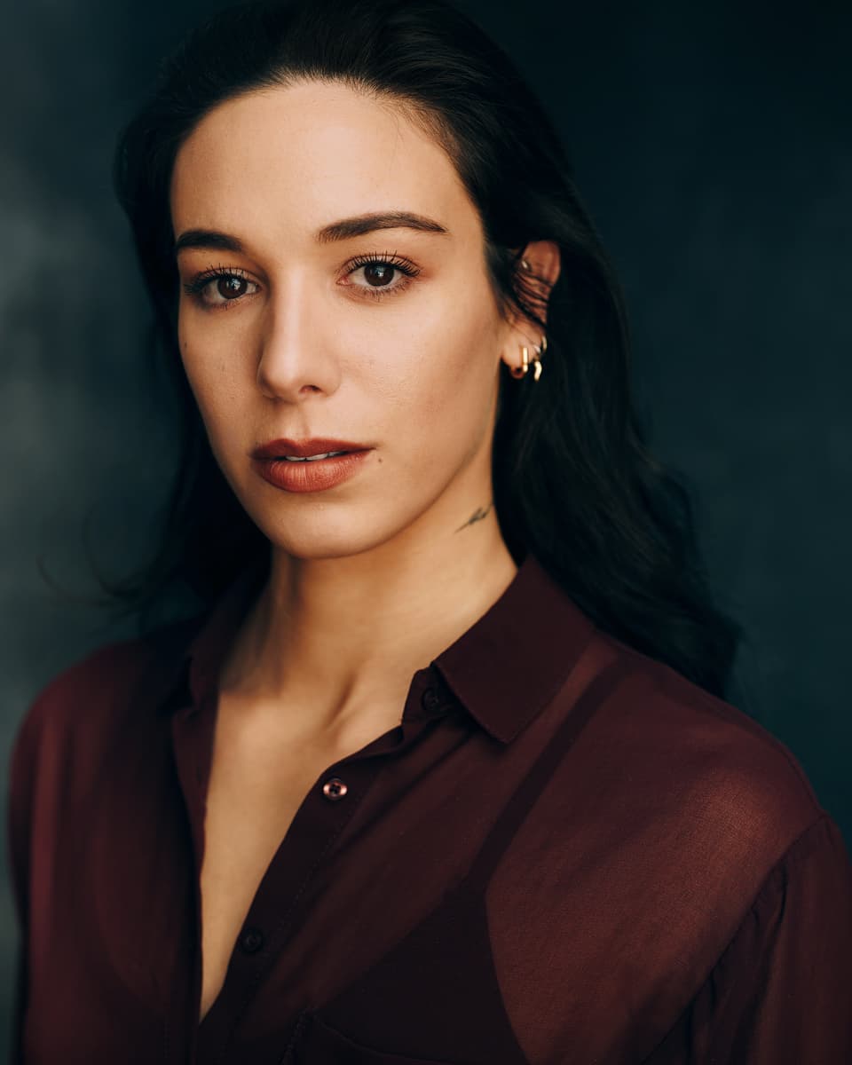 Dramatic actress headshot of a woman with long dark hair, layered gold earrings, and a small neck tattoo, wearing a sheer burgundy blouse with a steady and intense gaze against a deep teal studio background