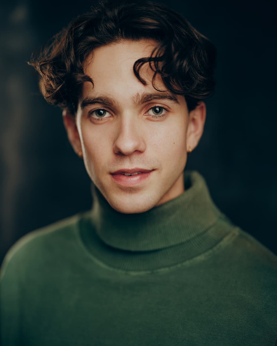 Editorial actor headshot of a young man with dark curly hair wearing a sage green turtleneck, with a soft natural expression and a small earring against a dark moody studio background