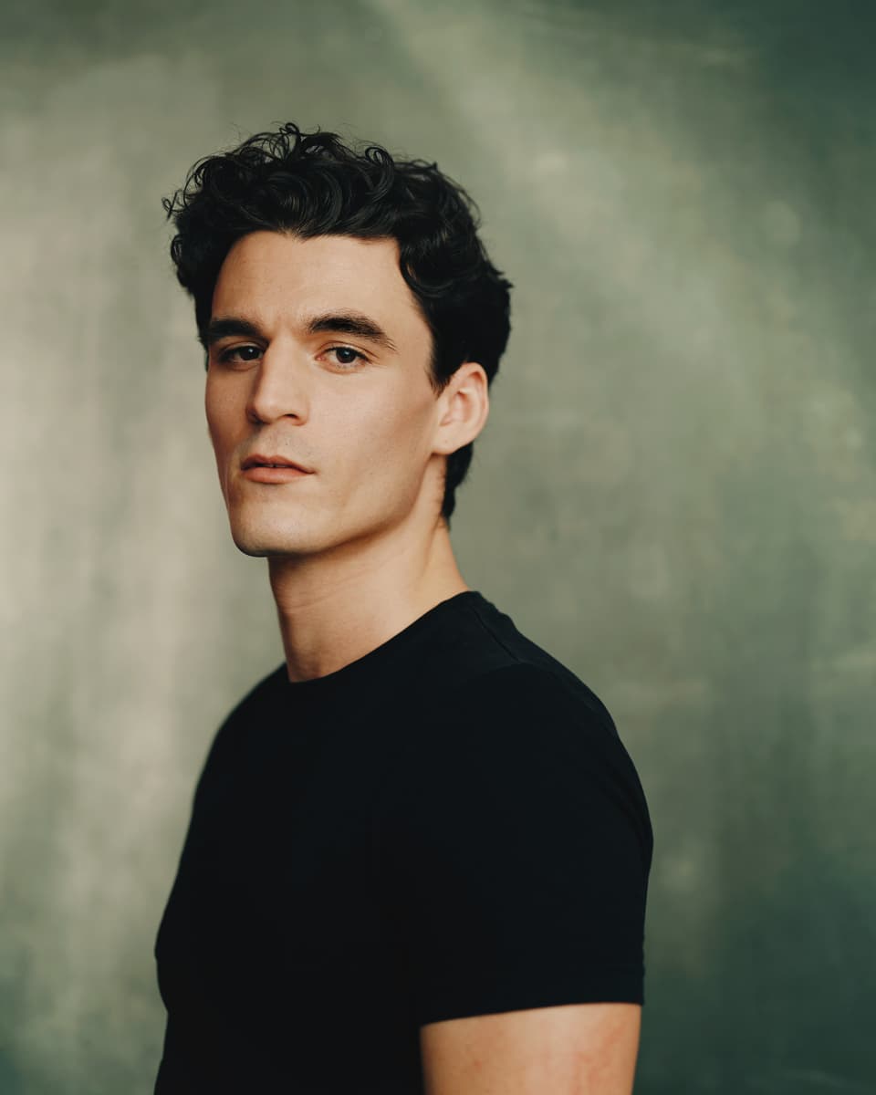 Actor headshot of a young man with dark wavy hair wearing a black t-shirt, looking over his shoulder with a calm, composed expression against a mottled sage-green studio backdrop