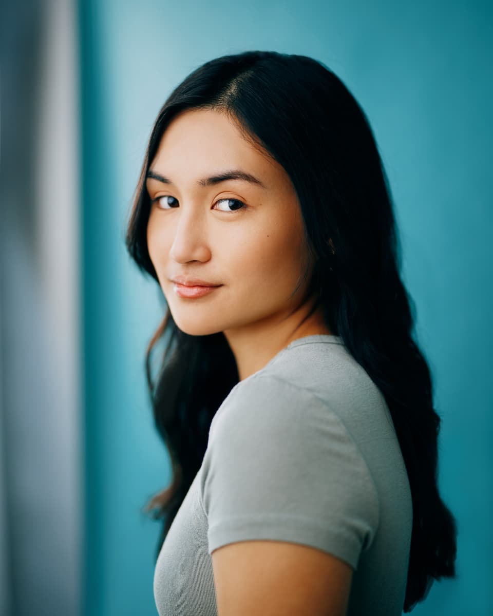 Actress headshot of a young woman with long dark hair glancing back over her shoulder with a soft smile, wearing a grey t-shirt against a bright teal studio backdrop