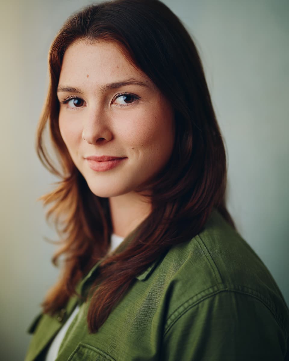 Theatrical actress headshot of a young woman with long auburn hair wearing a green jacket, with a soft approachable expression against a clean light grey studio background