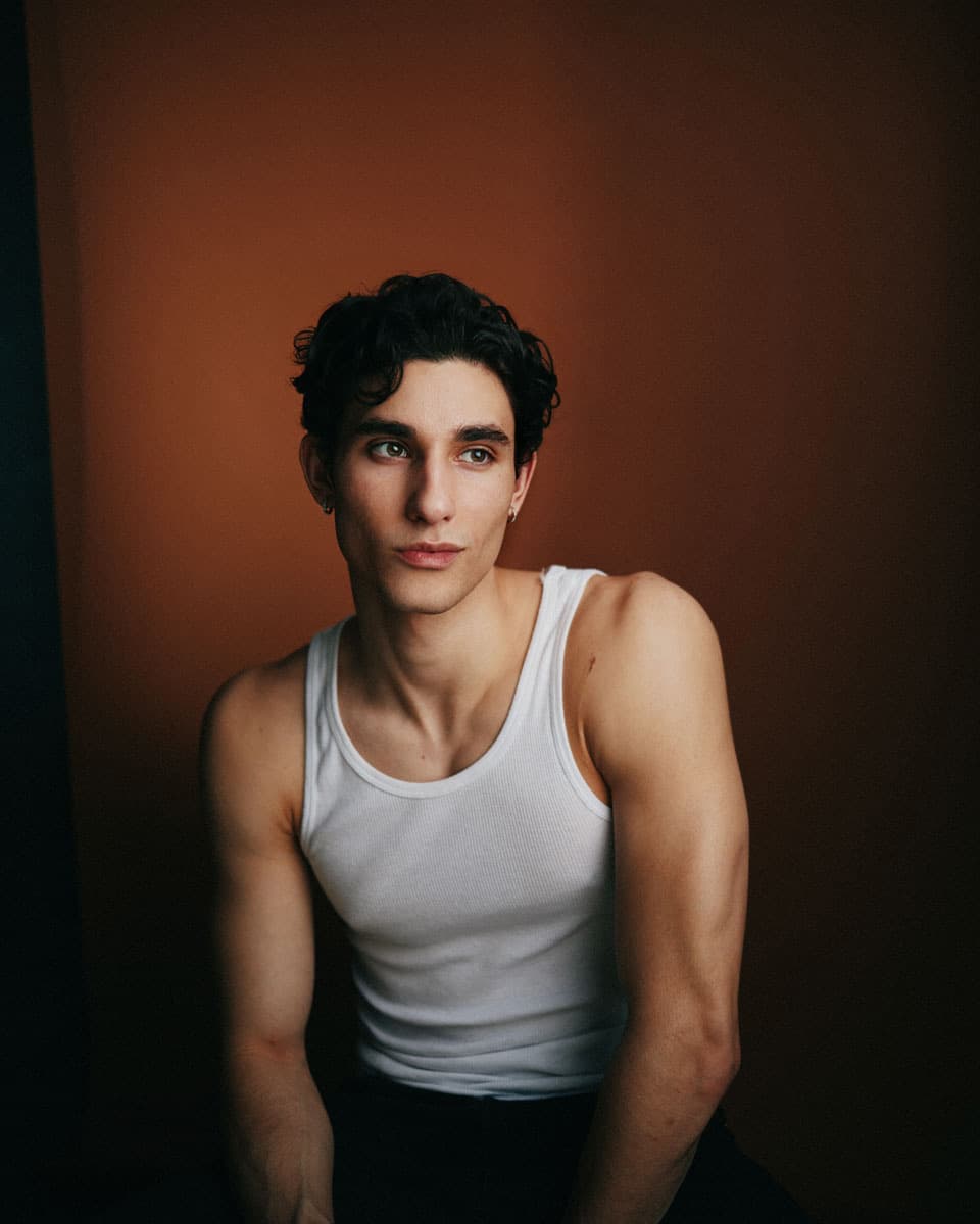 Theatrical actor headshot of a young man with dark curly hair, wearing a white tank top, with a calm direct gaze against a warm amber-brown backdrop