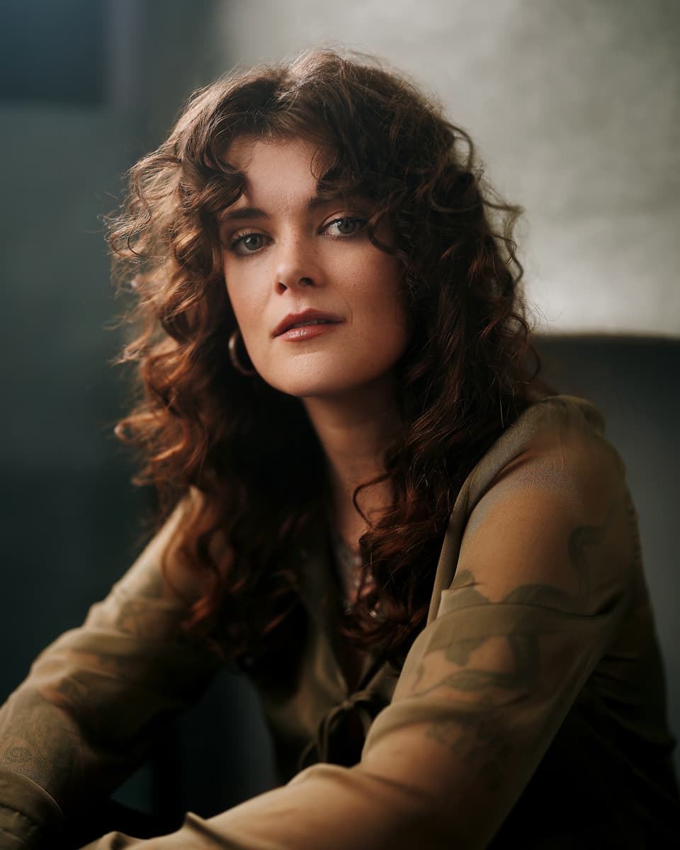 Theatrical headshot of a woman with voluminous dark curly hair wearing a sheer olive floral top, with a direct and expressive gaze against a warm brown studio backdrop