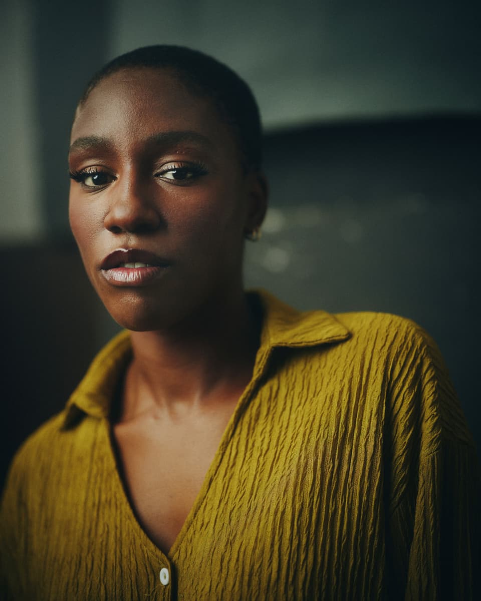 Toronto actor casting headshot of a woman with close-cropped hair wearing a mustard yellow textured shirt, with a calm and quietly intense expression against a dark charcoal-teal backdrop