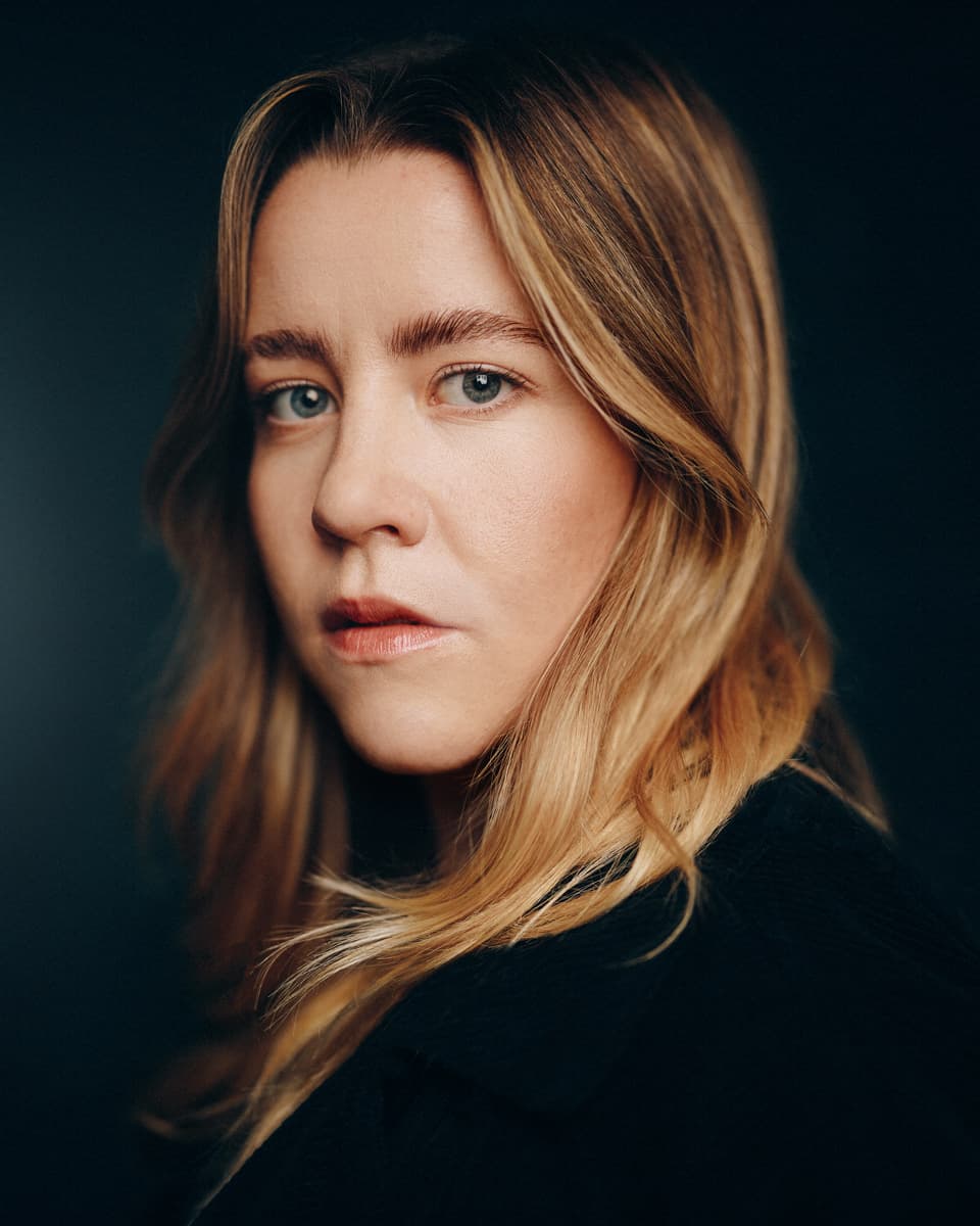 Toronto actor headshot of a woman with long blonde hair and blue eyes wearing a black top, with a composed and direct expression against a deep dark studio background
