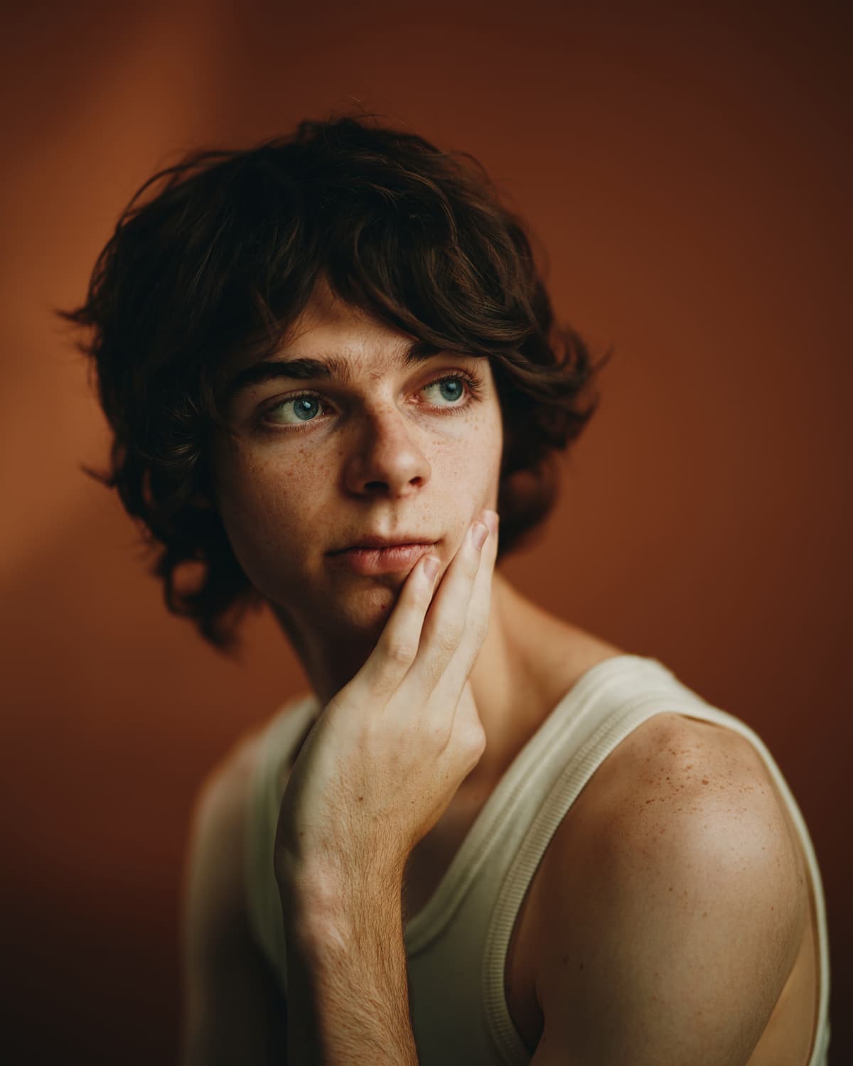Toronto actor headshot of a young man with curly dark hair and blue eyes, resting his hand against his cheek with a thoughtful, cinematic expression against a rich warm amber background