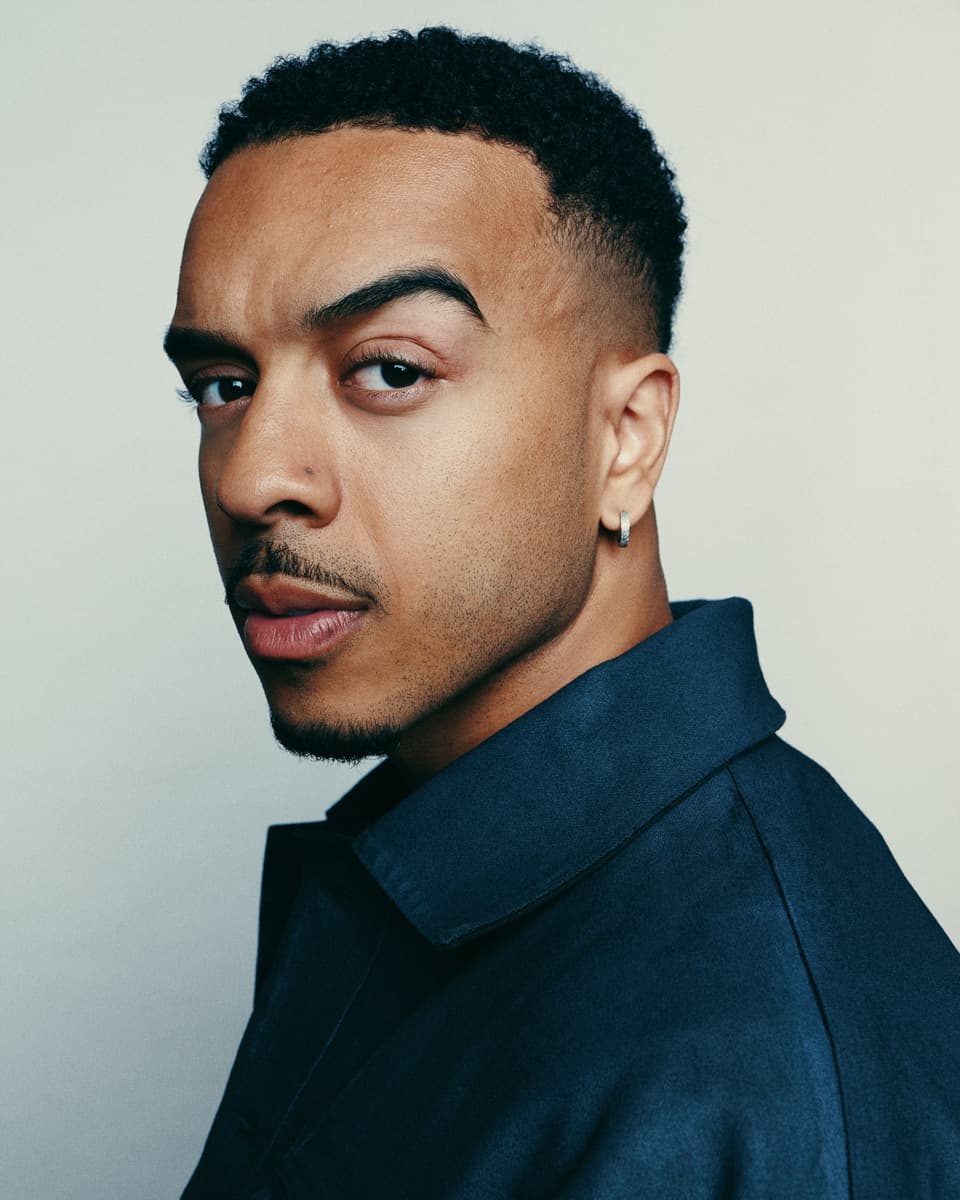 Toronto actor headshot of a man with short curly hair, a neat goatee, and a small hoop earring, wearing a dark navy shirt and looking back over his shoulder with a composed expression against a clean light grey background