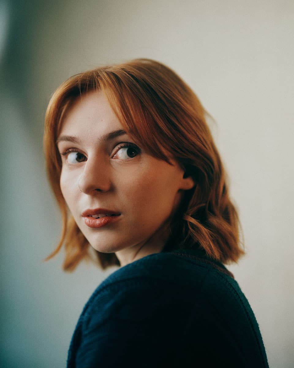 Toronto actor headshot of a young woman with short auburn hair and freckles, glancing back over her shoulder with a curious expression against a soft light background