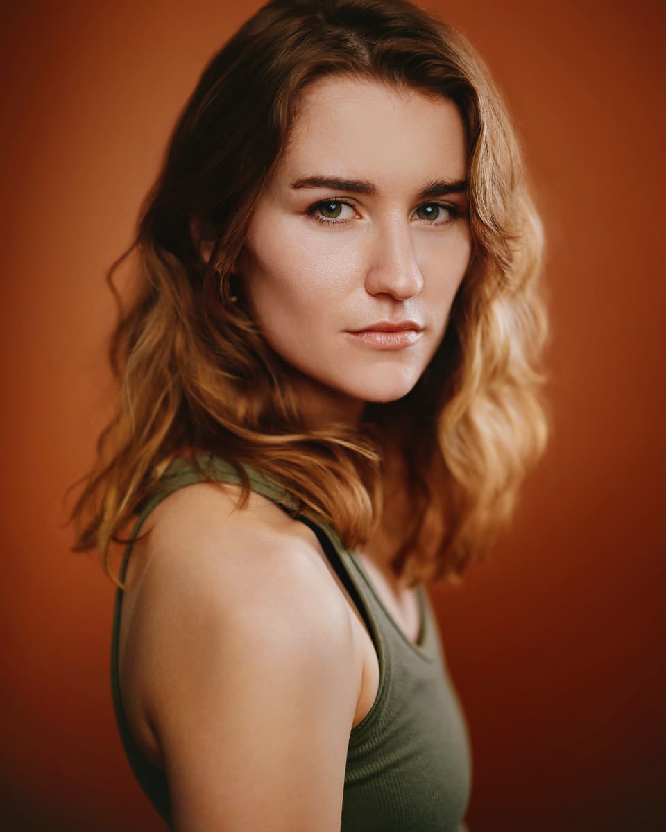 Toronto actor headshot of a young woman with wavy auburn hair wearing an olive green tank top, looking back over her shoulder with a direct, cool expression against a warm amber-red background
