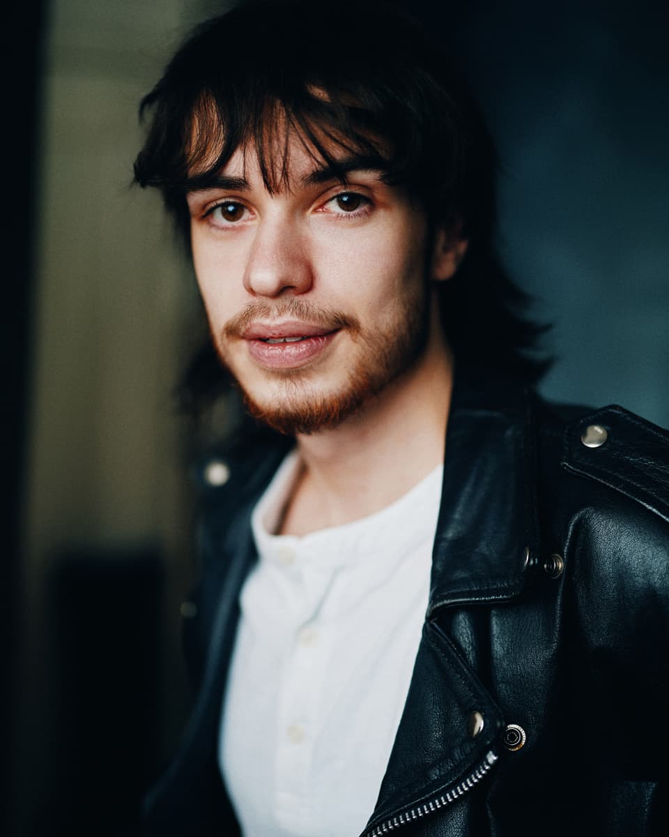 Toronto actor headshot of a young man with shaggy dark hair and a beard, wearing a white t-shirt under a black leather jacket, with a relaxed and natural expression against a dark moody background