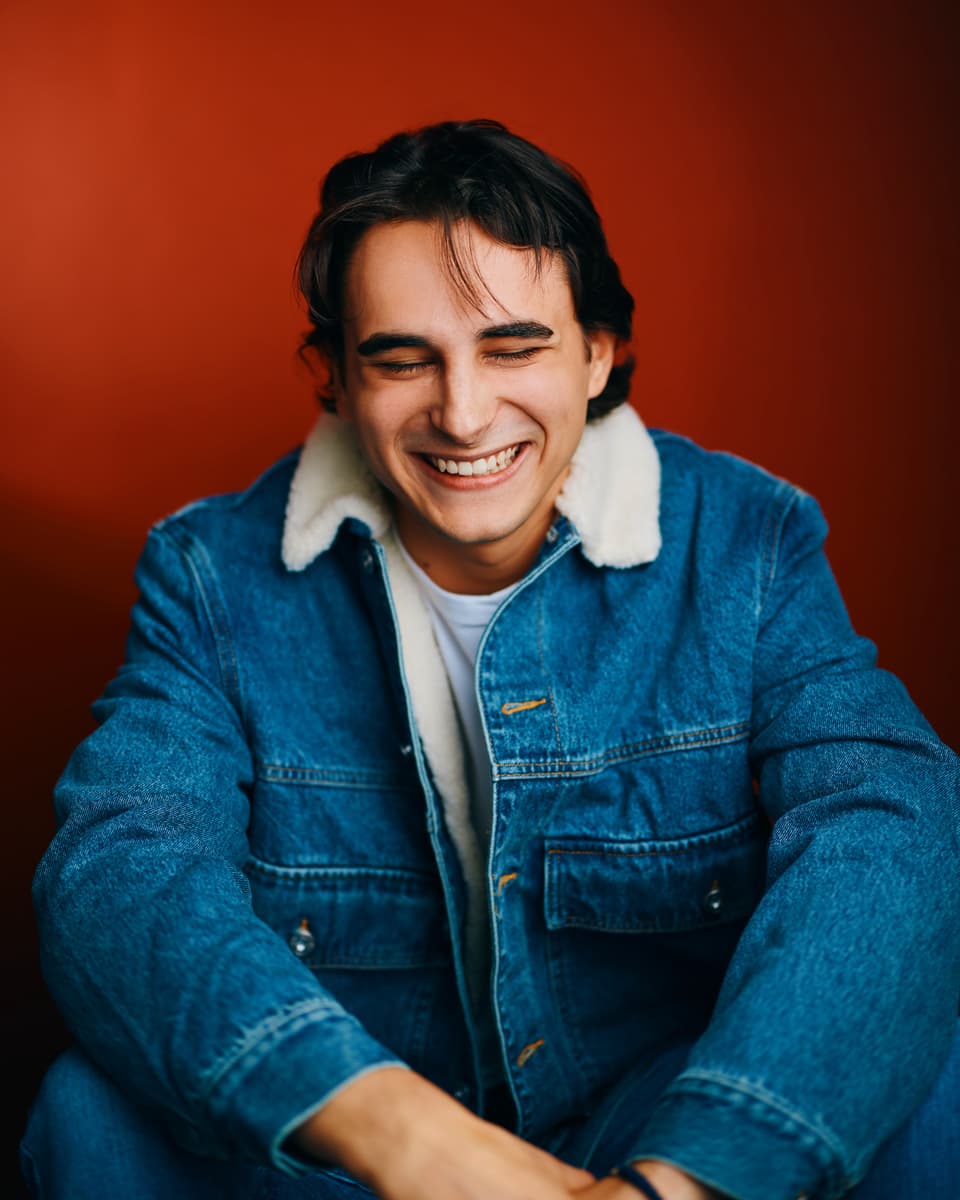 Toronto actor headshot of a young man with dark hair wearing a sherpa-collar denim jacket over a white t-shirt, laughing naturally with eyes closed against a bold warm amber-red background