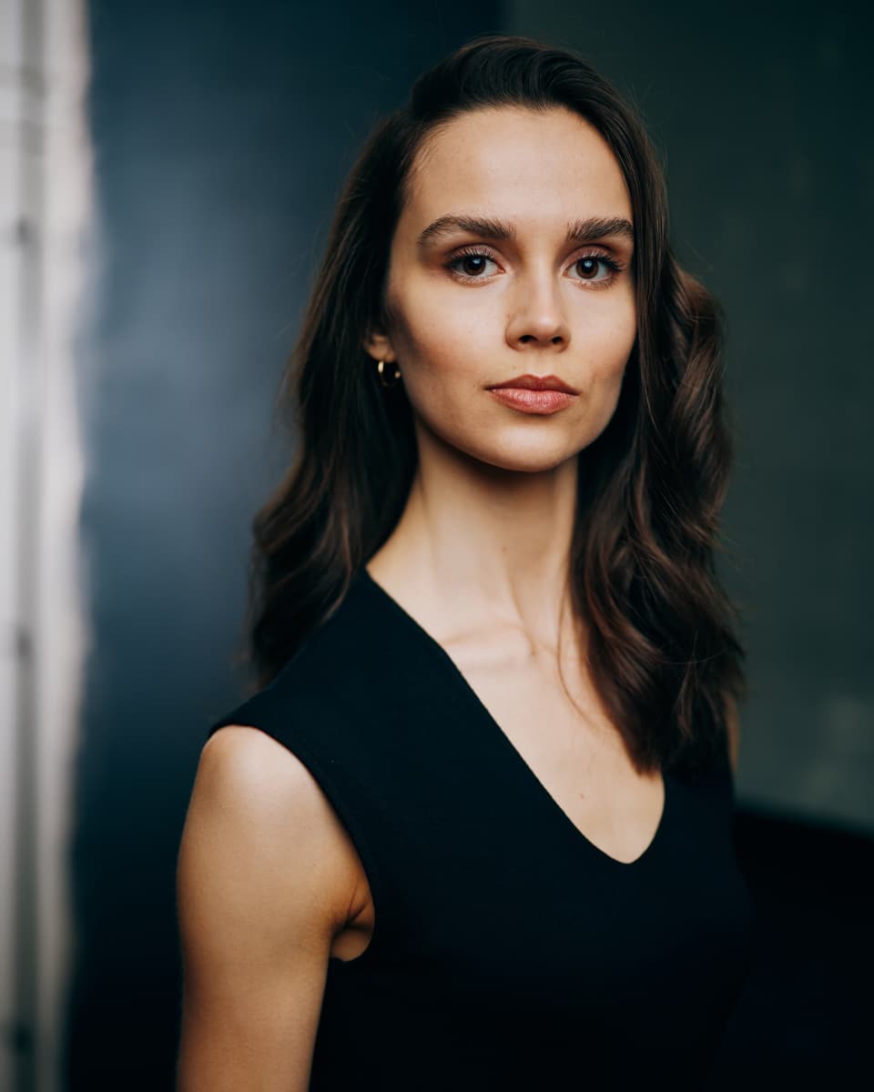 Toronto film actor headshot of a woman with long dark wavy hair wearing a black sleeveless V-neck top, with bold brows and a composed expression against a dark cinematic teal backdrop