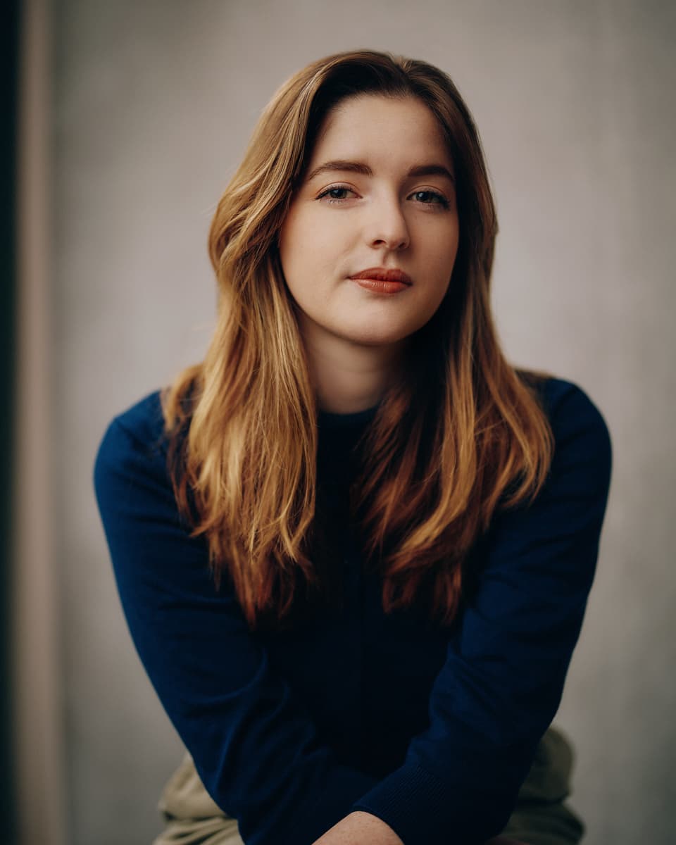 Toronto singer headshot of a young woman with long auburn hair wearing a deep blue long-sleeve top, seated with hands clasped and a calm, direct expression against a soft warm grey studio background