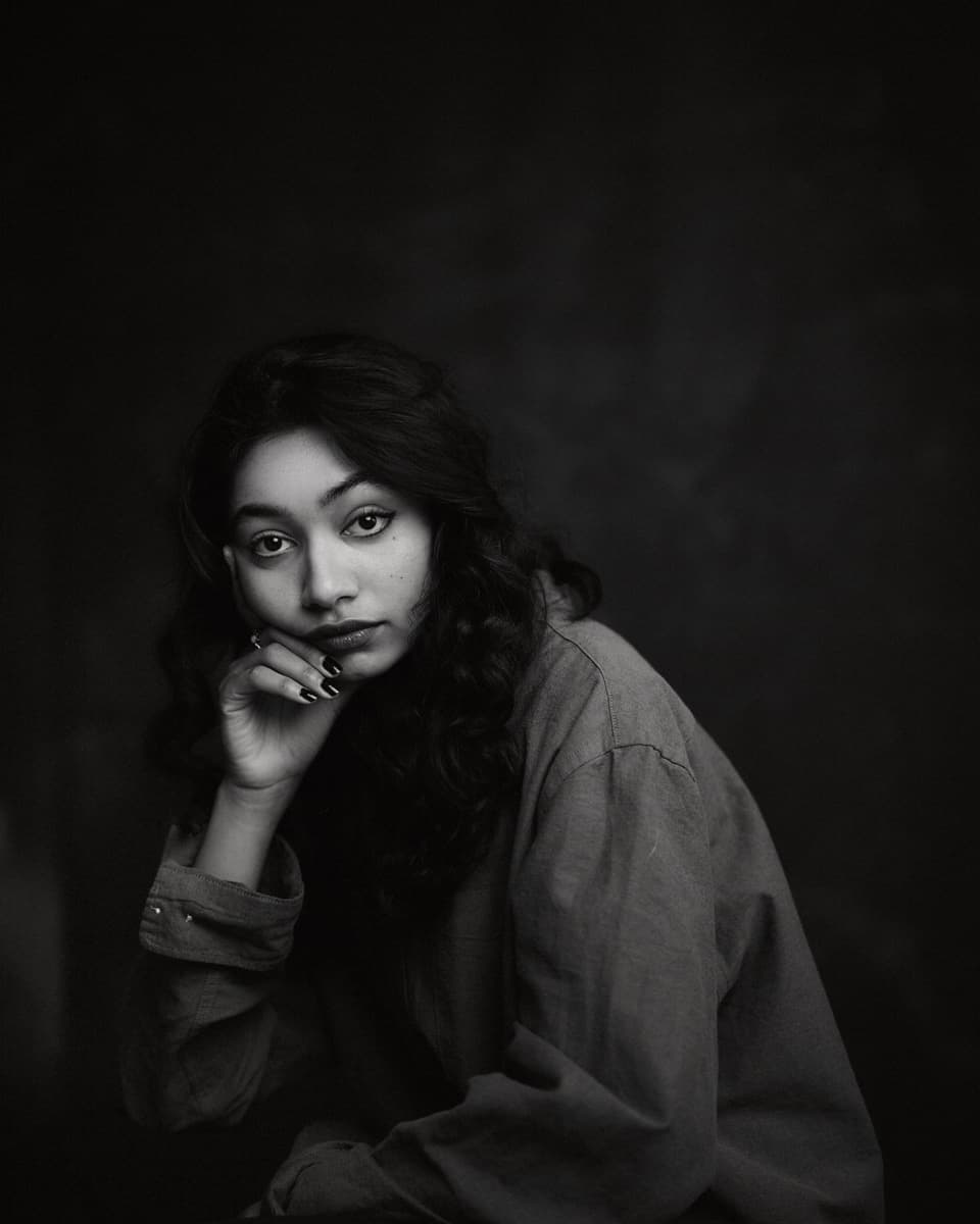 Toronto theatre actor headshot of a young woman with long dark wavy hair, resting her chin on her dark-painted hand and gazing directly at the camera with a pensive expression, shot in black and white against a deep dark background