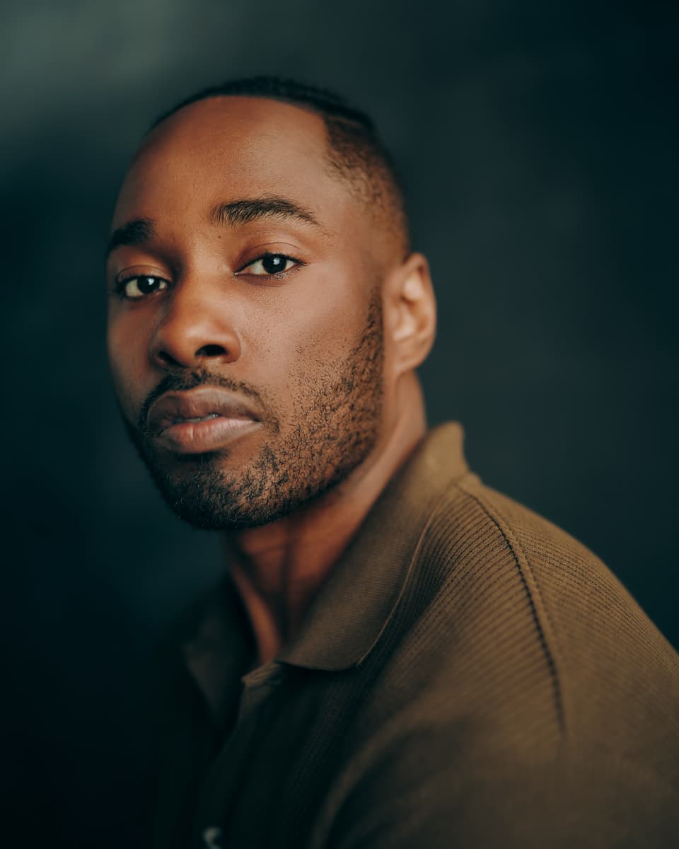 Toronto voice actor headshot of a man with short hair wearing an olive ribbed polo, with a strong, steady gaze against a dark teal studio background