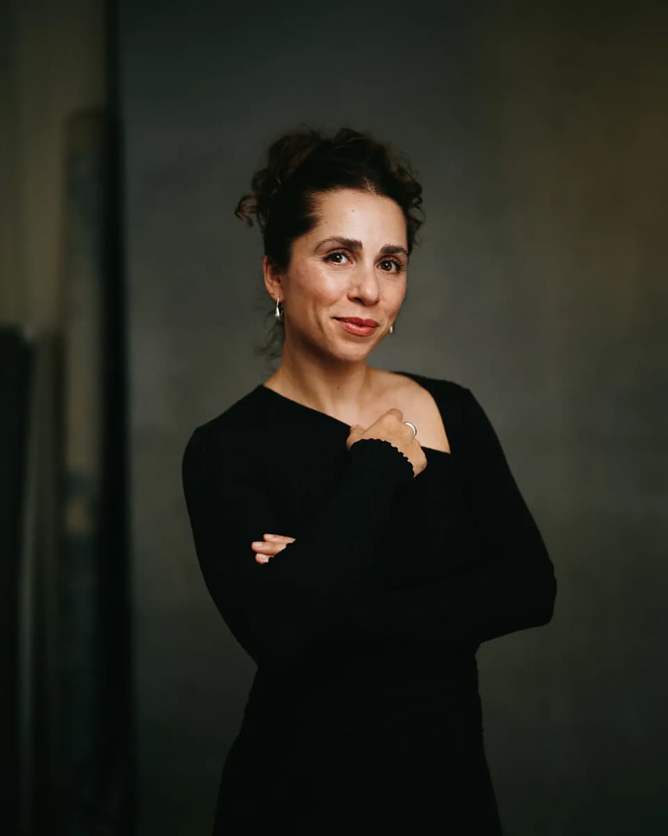 Gabriella Pirraglia, associate photographer at Calvin Thomas Studio, wearing a black top with arms crossed and a warm expression against a moody grey-green studio background