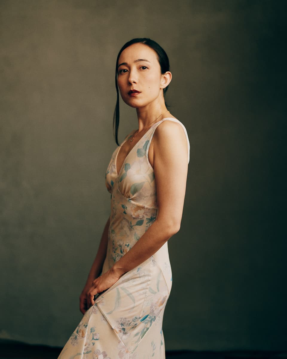Toronto actor headshot of a woman with dark hair pulled back, wearing a sheer floral slip dress, standing in three-quarter profile with a composed and striking gaze against a moody olive-grey studio backdrop