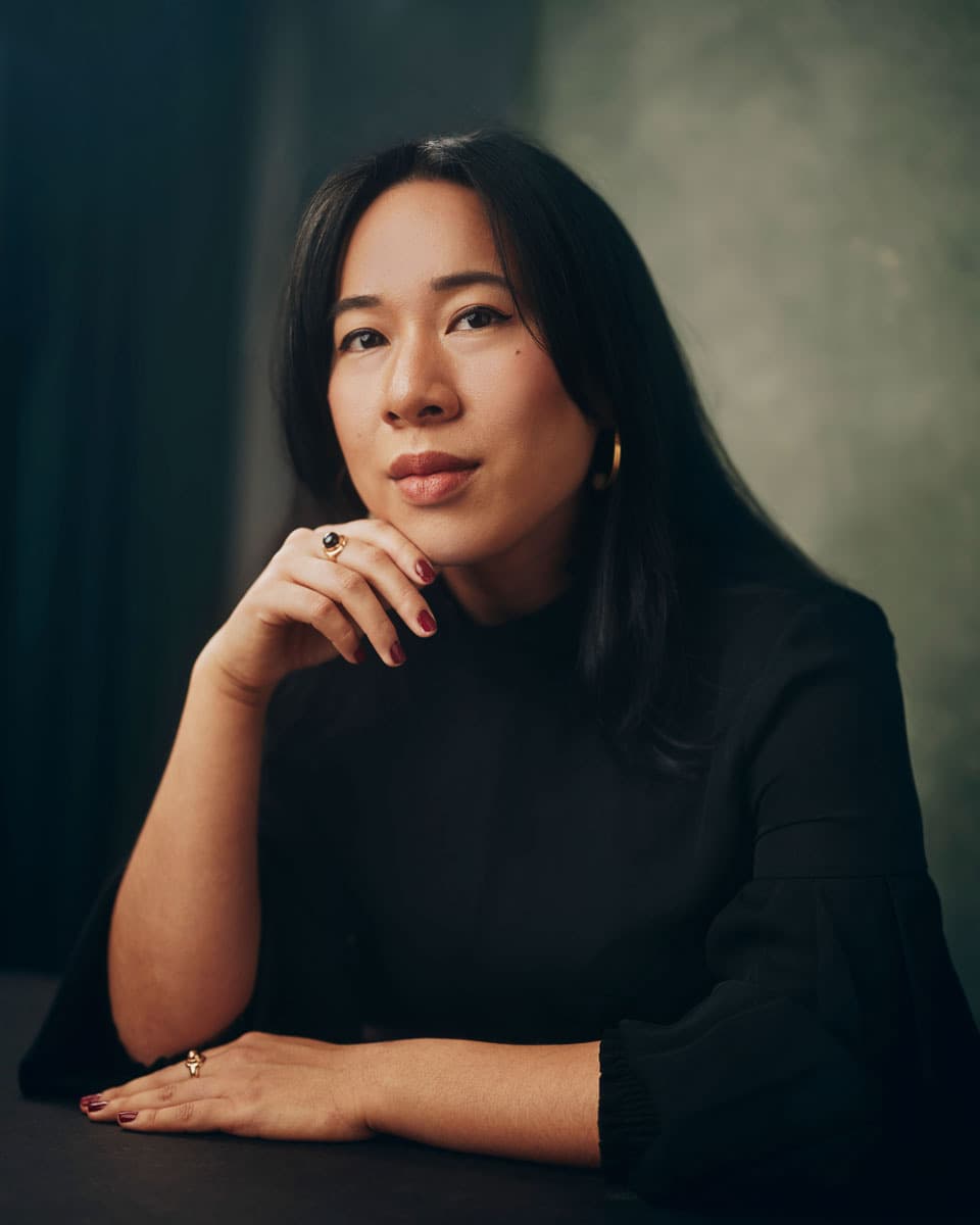 Toronto professional headshot of a woman with long dark hair wearing a black top, resting her chin on her hand with gold rings and red nails, with a composed and confident expression against a dark teal studio background