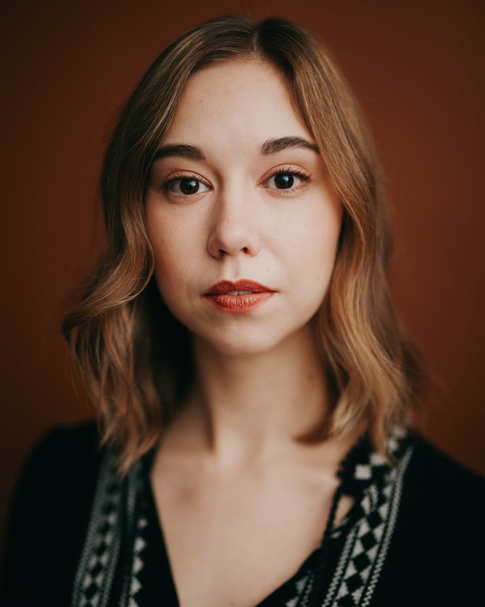 Toronto actor headshot of a woman with long dark wavy hair in a soft grey oversized sweater, one hand lightly touching her collarbone, with a quietly intense gaze against a moody dark green-grey background