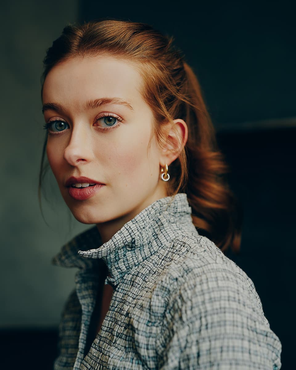 Actor casting headshot of a young woman with auburn hair swept back, blue-grey eyes, and gold hoop earrings, wearing a light grey plaid shirt and looking over her shoulder with a calm, neutral expression against a dark moody background