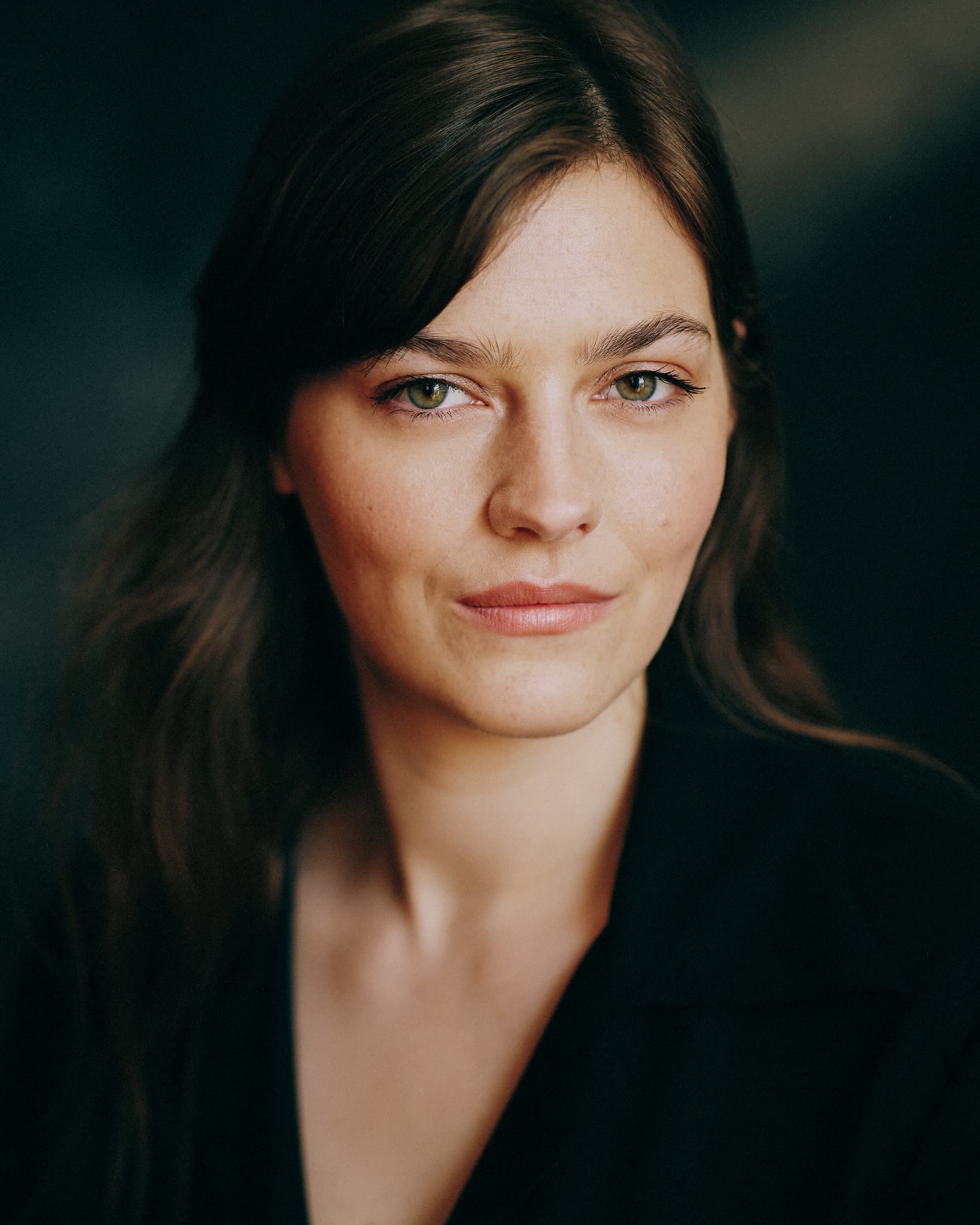 Toronto actor headshot of a young woman with long dark brown hair in a black V-neck top, gazing directly at the camera against a deep dark teal background