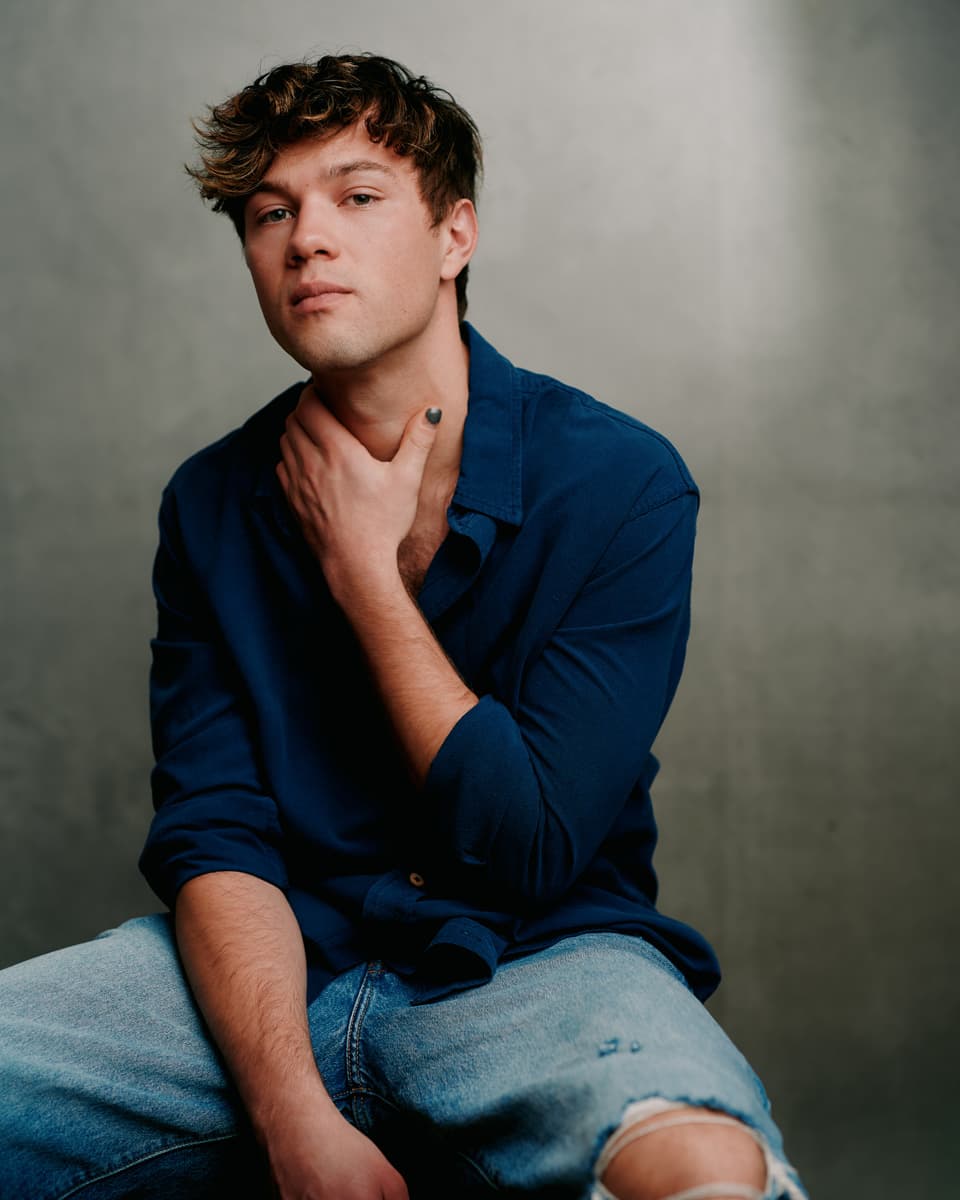 Theatrical headshot of a young man with tousled curly brown hair and green eyes, cradling his face in both hands to reveal dark-painted nails, wearing a blue tie-dye shirt, with an introspective expression against a pale grey background