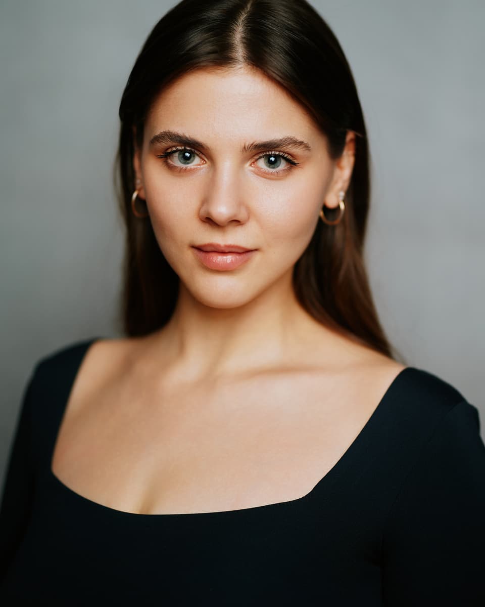 Toronto actress headshot with natural makeup — a young woman with long straight dark hair and blue-grey eyes in a black square-neck top against a clean light grey backdrop