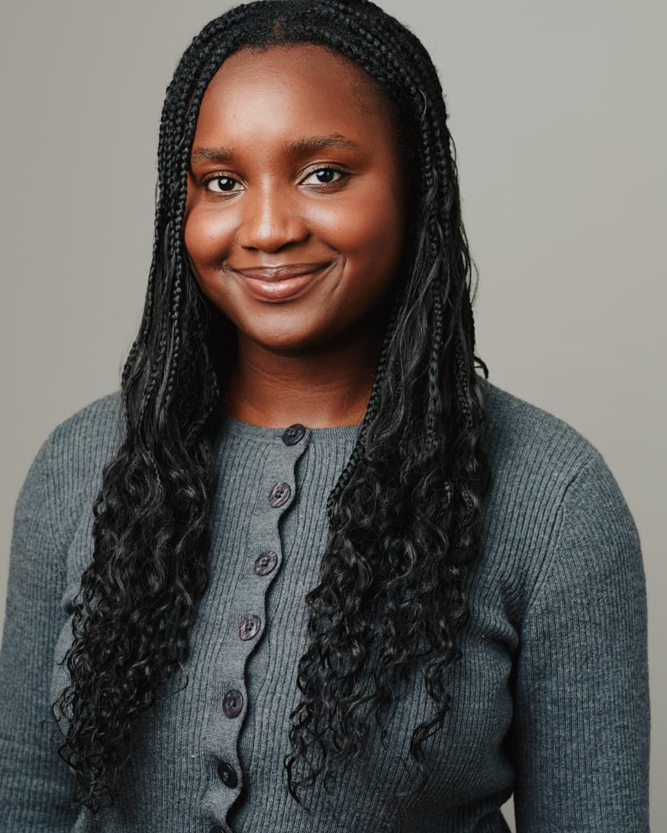 Toronto performing artist headshot of a young woman with long dark wavy hair, wearing a terracotta button-up shirt over a white top, beaming with a wide natural smile against a soft warm grey studio background