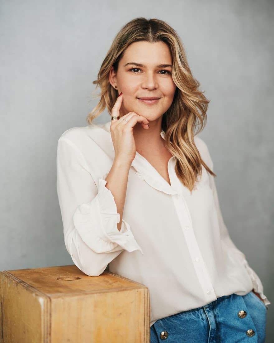 Toronto business headshot of a woman with wavy blonde hair in a white ruffled blouse, resting her chin on her hand with a warm smile against a soft grey studio background