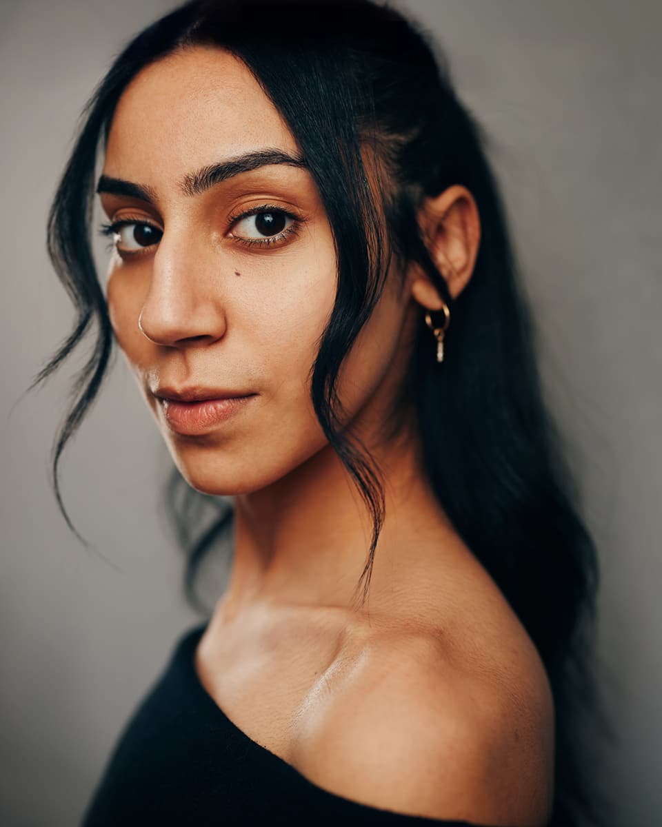 Theatrical studio headshot of a young woman with long dark hair, a nose ring, and small gold hoop earrings, wearing an off-shoulder black top, gazing directly at the camera with an intense, quietly confident expression against a warm grey background
