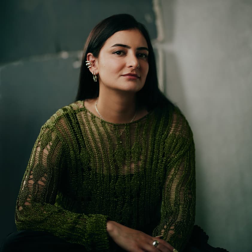 Toronto editorial portrait of a young woman with straight black hair in a textured green lace knit sweater and gold ear cuffs, seated against a raw concrete wall