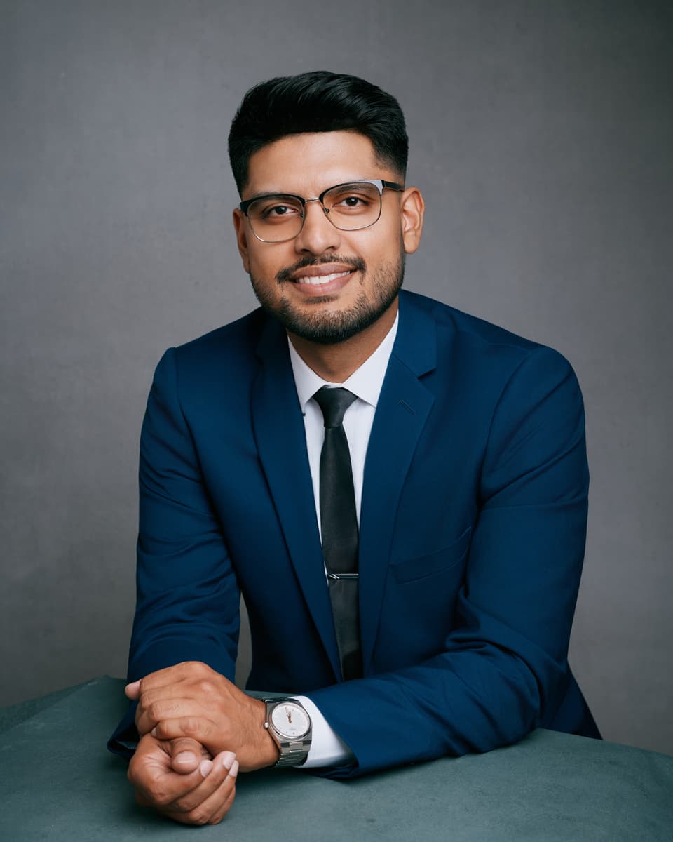 Toronto corporate headshot of a man in a navy suit, white dress shirt, black tie, and rectangular glasses, smiling warmly with hands clasped and a silver watch visible against a dark grey studio background
