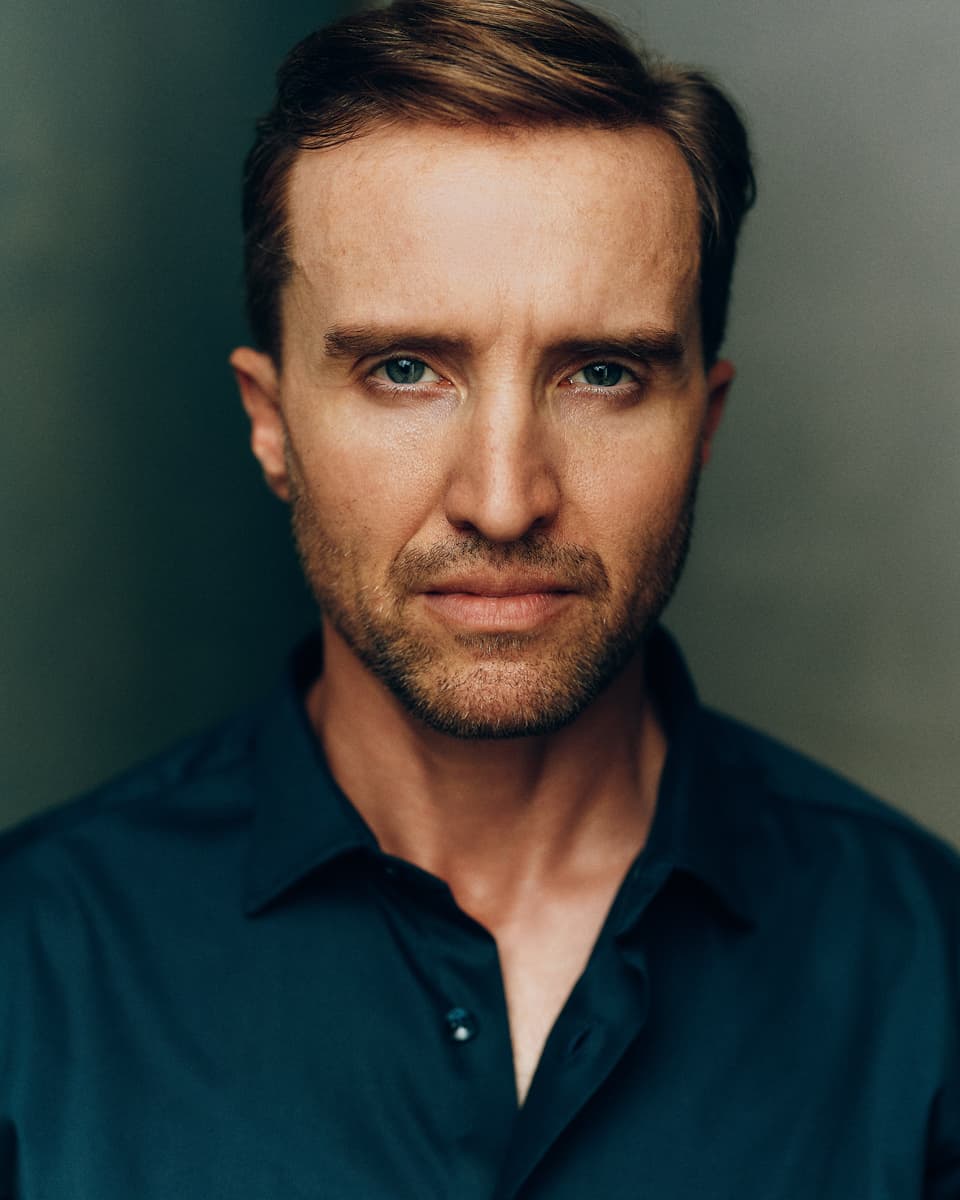 Cinematic Toronto film actor headshot of a man with short sandy hair and blue eyes, with a strong direct gaze against a gradient blue-grey background