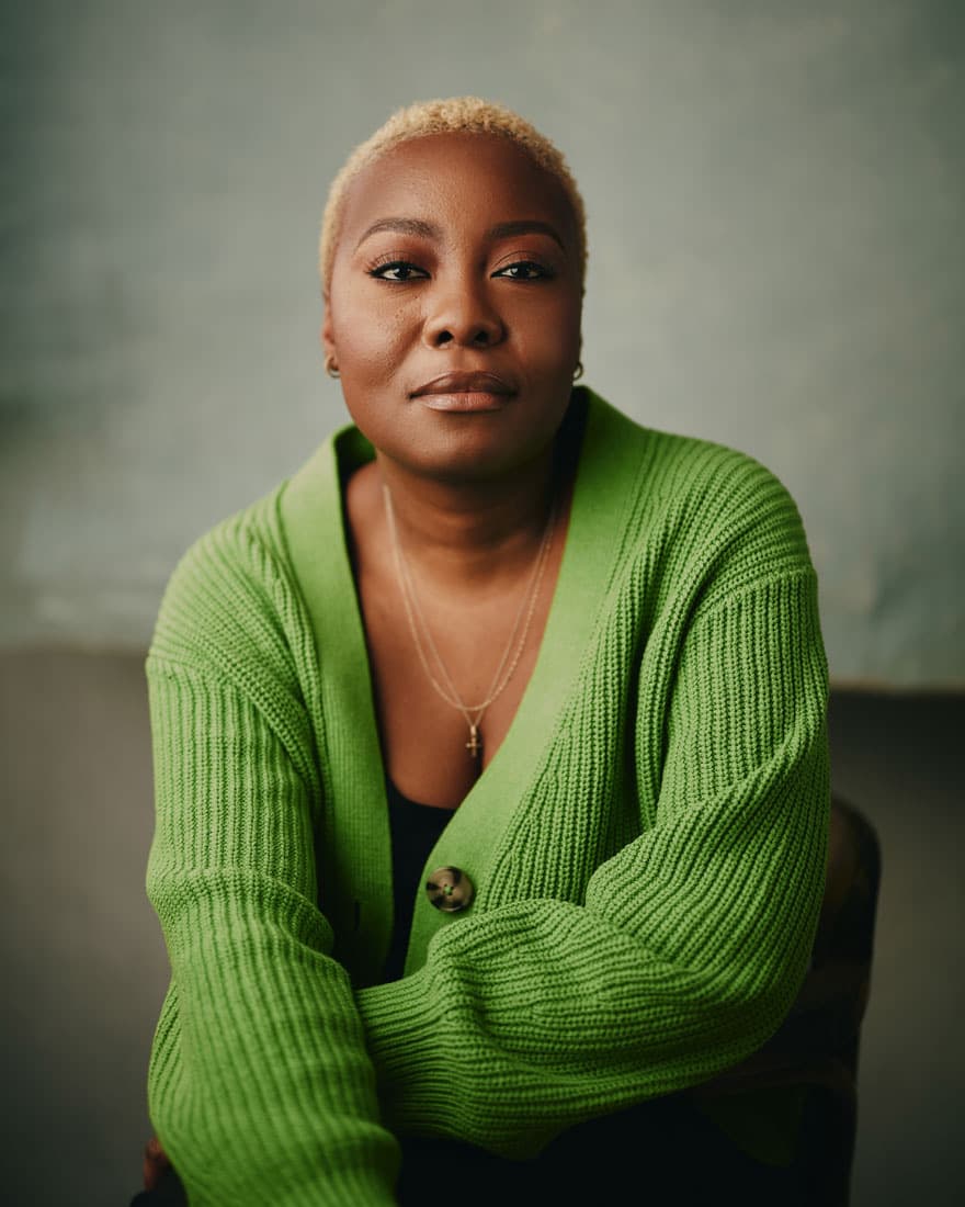 Toronto musician studio headshot of a woman with short blonde natural hair, wearing a bold green chunky knit cardigan and a delicate gold necklace, arms folded with a calm, self-assured gaze against a textured grey-green background